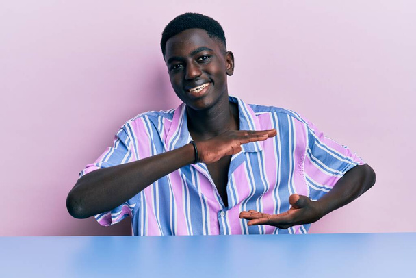 Nuori afrikkalainen amerikkalainen mies päällään rento vaatteita istuu pöydällä eleiden kädet osoittavat iso ja suuri koko merkki, mitta symboli. hymyillen katsoen kameraan. mittauskäsite.  - Valokuva, kuva