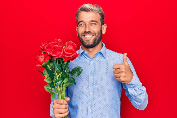 Jeune homme romantique blond beau tenant bouquet de belles roses sur fond rouge souriant heureux et positif, pouce levé faire excellent et signe d'approbation - Photo, image