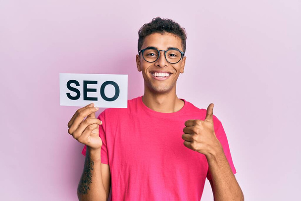 Young handsome african american man holding seo message paper smiling happy and positive, thumb up doing excellent and approval sign  - Photo, Image