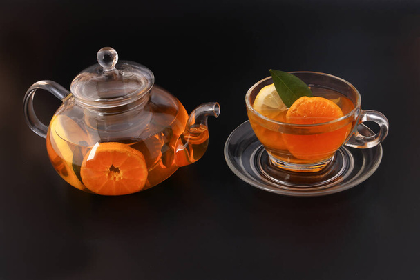 Liquido tè limone arancia fetta foglia verde cannella bastone in vetro trasparente tazza da tè piattino teiera bollitore su sfondo nero - Foto, immagini