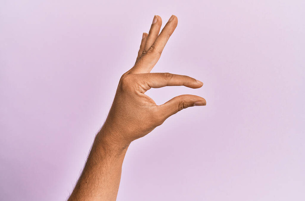 Рука и рука кавказского молодого человека на розовом изолированном фоне, собирающего и захватывающего невидимую вещь, держащего объект пальцами, показывающими пространство  - Фото, изображение