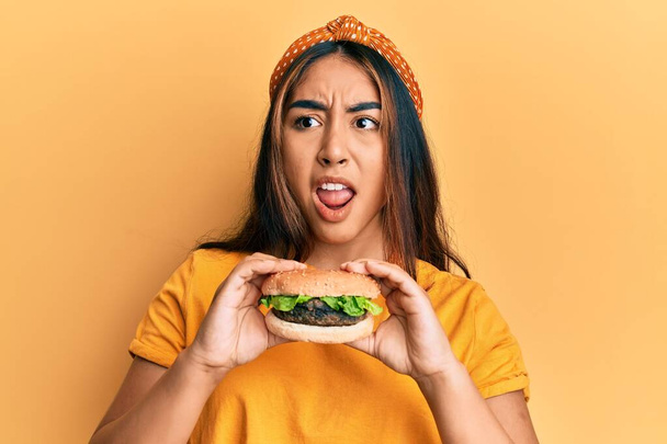 Junge lateinamerikanische Frau isst einen leckeren klassischen Burger wütend und verrückt schreit frustriert und wütend, schreit vor Wut. Wut und aggressives Konzept.  - Foto, Bild