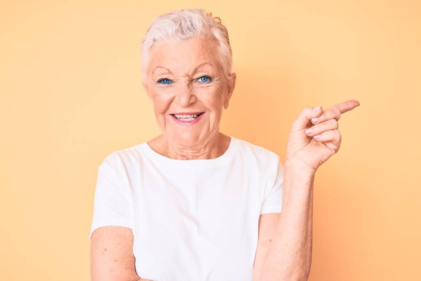 Belle femme âgée aux yeux bleus et aux cheveux gris portant un t-shirt blanc classique sur fond jaune avec un grand sourire sur le visage, pointant avec la main et le doigt vers le côté en regardant la caméra.  - Photo, image