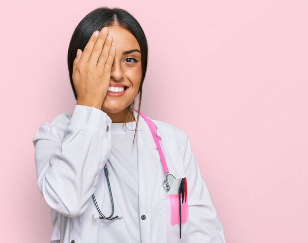 Schöne hispanische Frau in Arztuniform und Stethoskop, die ein Auge mit der Hand bedeckt, selbstbewusstes Lächeln im Gesicht und überraschende Emotionen.  - Foto, Bild