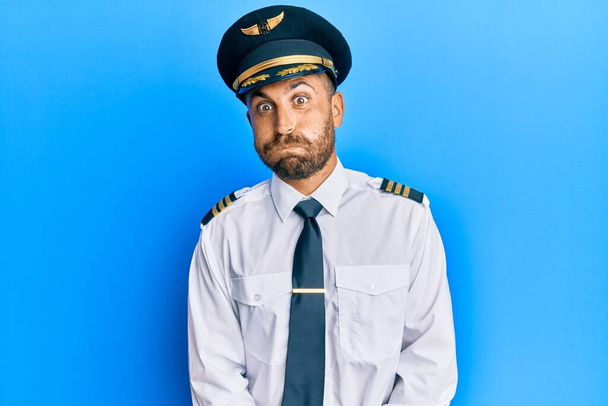Handsome man with beard wearing airplane pilot uniform puffing cheeks with funny face. mouth inflated with air, crazy expression.  - Foto, afbeelding