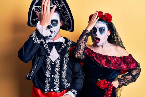 Young couple wearing mexican day of the dead costume over yellow surprised with hand on head for mistake, remember error. forgot, bad memory concept.  - Photo, Image