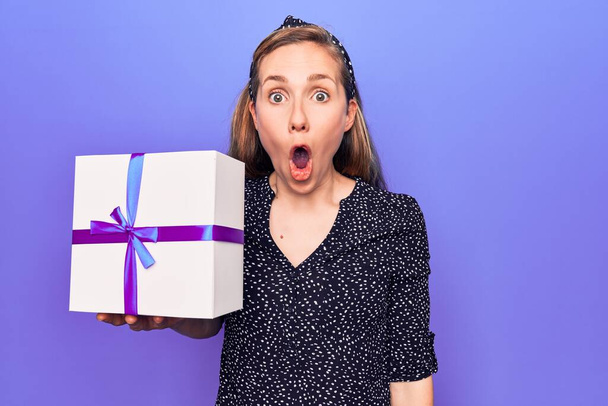 Jeune belle femme blonde tenant boîte cadeau d'anniversaire effrayé et étonné avec bouche ouverte pour la surprise, visage incrédule  - Photo, image