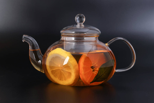 Рідкий чай лимонно-помаранчевий шматочок зеленого листя кориці паличка в прозорому скляному чайнику на чорному фоні
 - Фото, зображення