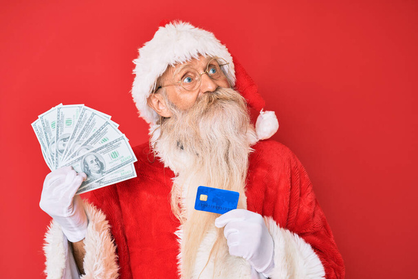 Staruszek w kostiumie Świętego Mikołaja trzymający dolary i kartę kredytową uśmiechający się patrząc w bok i patrząc myśląc.  - Zdjęcie, obraz