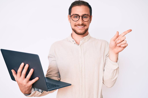 Genç yakışıklı adam dizüstü bilgisayarını tutuyor gülümsüyor mutlu bir şekilde elini ve parmağını yana doğru gösteriyor.  - Fotoğraf, Görsel