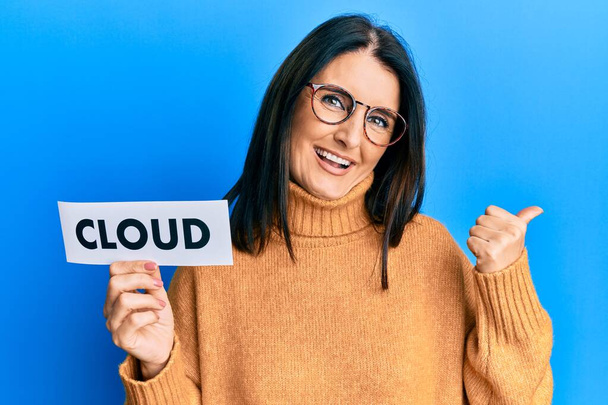 Middle age brunette woman holding paper with cloud word pointing thumb up to the side smiling happy with open mouth  - Φωτογραφία, εικόνα