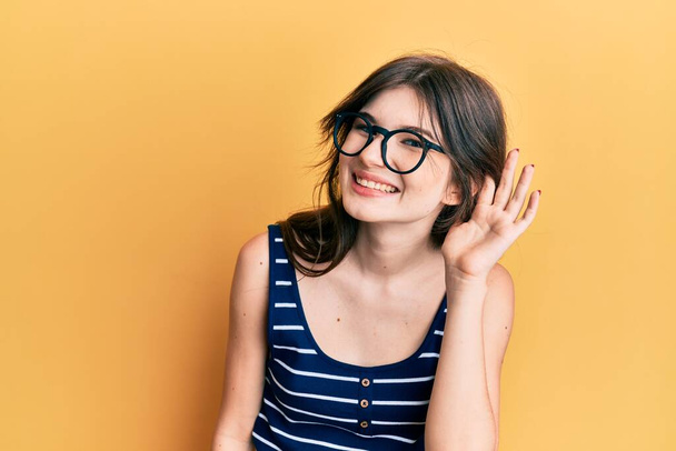 Young beautiful caucasian girl wearing casual clothes and glasses smiling with hand over ear listening and hearing to rumor or gossip. deafness concept.  - Photo, Image