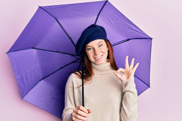 若いです赤い頭の女の子保持紫色の傘を身に着けています新鮮なベレー帽を身に着けています指でokサイン,笑顔優しいジェスチャー優れたシンボル  - 写真・画像