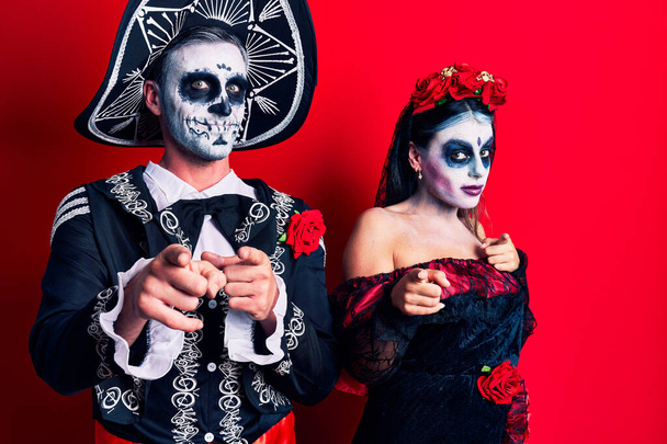 Jong stel draagt Mexicaanse dag van het dode kostuum over rode wijzende vingers naar camera met vrolijk en grappig gezicht. goede energie en vibes.  - Foto, afbeelding