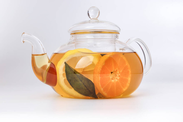 Folyékony tea citrom narancs szelet zöld levél fahéj bot átlátszó üveg teáskanna vízforraló fehér alapon - Fotó, kép