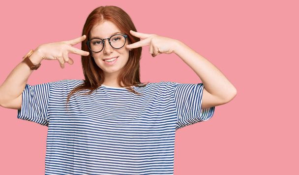 Young read head woman wearing casual clothes and glasses doing peace symbol with fingers over face, smiling cheerful showing victory  - Fotoğraf, Görsel