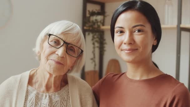 Genç Asyalı kadın bakıcı ve yaşlı beyaz kadının kamera önünde birlikte poz verirken ve evde gülümserken göğsünü dik tut. - Video, Çekim