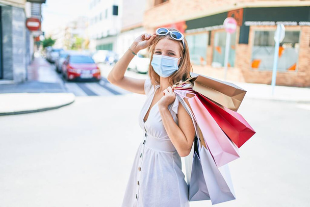 Jeune femme blonde portant un masque médical tenant des sacs à provisions dans la rue de la ville - Photo, image