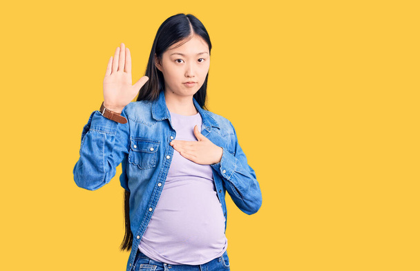 Jovem mulher chinesa bonita grávida esperando bebê jurando com a mão no peito e palma aberta, fazendo uma promessa de lealdade juramento  - Foto, Imagem