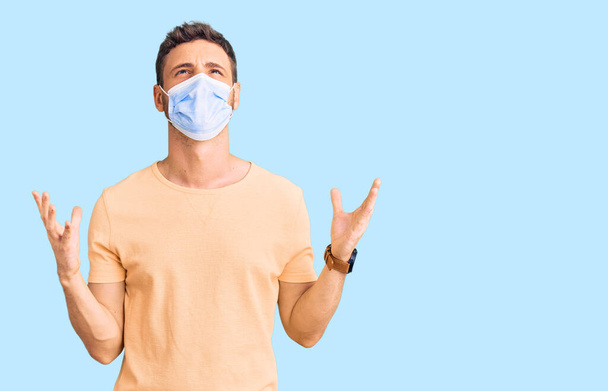 Handsome young man with bear wearing medical mask for coronavirus crazy and mad shouting and yelling with aggressive expression and arms raised. frustration concept.  - Foto, imagen