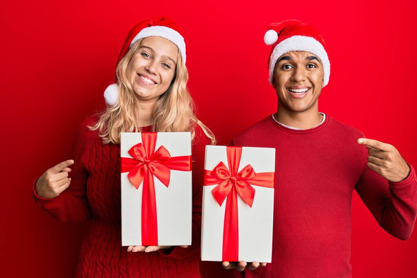 Νεαρό διαφυλετικό ζευγάρι φοράει χριστουγεννιάτικο καπέλο και κρατώντας ένα δώρο χαμογελώντας χαρούμενος δείχνοντας με το χέρι και το δάχτυλο  - Φωτογραφία, εικόνα