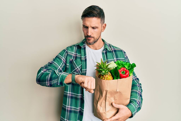 Schöner Mann mit Bart hält Papiertüte mit Lebensmitteln in der Hand und kontrolliert die Uhrzeit an der Armbanduhr, entspannt und selbstbewusst  - Foto, Bild