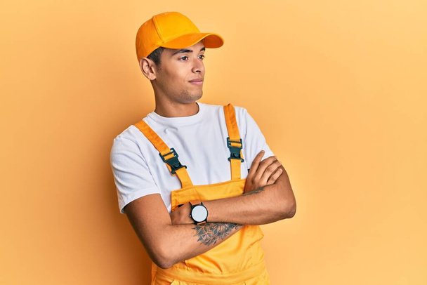 Giovane bell'uomo afroamericano che indossa tuttofare uniforme su sfondo giallo guardando a lato con le braccia incrociate convinto e fiducioso  - Foto, immagini