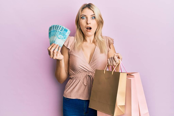 Young caucasian woman holding brazil real banknotes and shopping bags afraid and shocked with surprise and amazed expression, fear and excited face.  - Foto, imagen