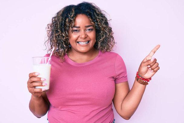 jong Afrikaans amerikaans plus size vrouw drinken glas melk lachend gelukkig wijzend met hand en vinger naar de zijkant  - Foto, afbeelding
