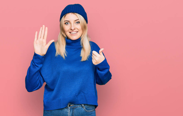 Молода біла жінка носить вовняний зимовий светр і кепку, показуючи і вказуючи пальцями номер шість, посміхаючись впевнено і щасливо
.  - Фото, зображення