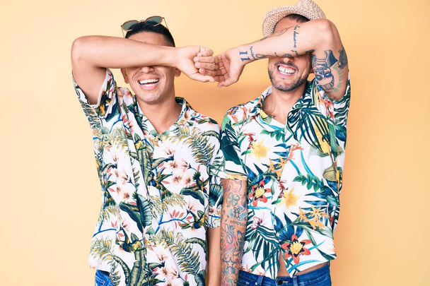Jeune couple gay de deux hommes portant un chapeau d'été et chemise hawaïenne couvrant les yeux avec le bras souriant gai et drôle. concept aveugle.  - Photo, image