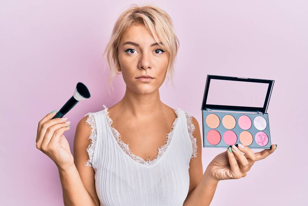 Young blonde girl holding makeup brush and blush relaxed with serious expression on face. simple and natural looking at the camera.  - Photo, Image