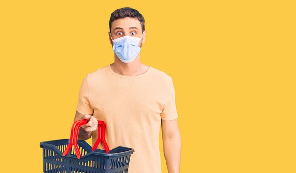Handsome young man with bear wearing shopping basket and medical mask scared and amazed with open mouth for surprise, disbelief face  - Foto, Imagen