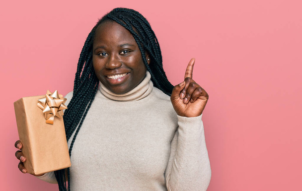 Young black woman with braids holding gift surprised with an idea or question pointing finger with happy face, number one  - Photo, image