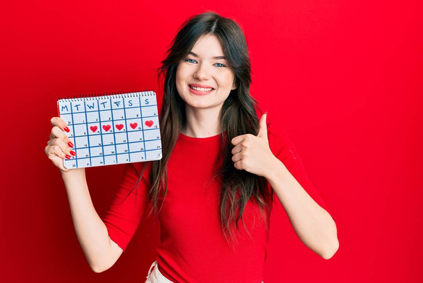 Young beautiful caucasian girl holding heart calendar smiling happy and positive, thumb up doing excellent and approval sign  - Photo, Image