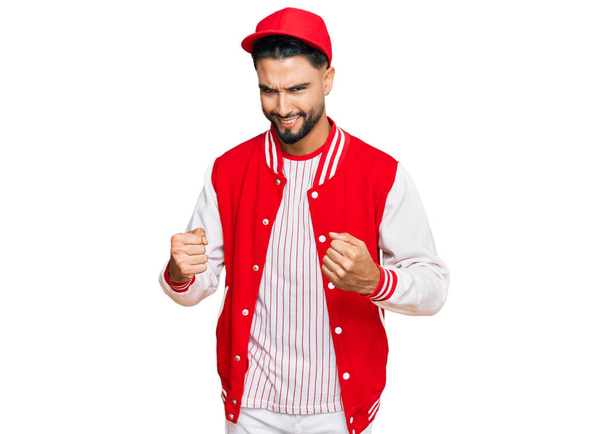 Junger Mann mit Bart in Baseball-Uniform fiebert mit erhobenen Armen und geschlossenen Augen dem Erfolg entgegen und feiert lächelnd. Siegerkonzept.  - Foto, Bild