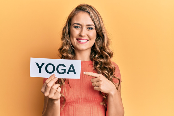 Giovane ragazza bionda in possesso di carta messaggio yoga sorridente felice indicando con mano e dito  - Foto, immagini