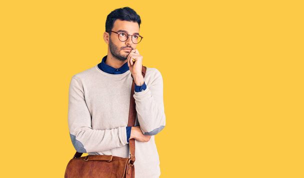 Young handsome hispanic man wearing leather bag serious face thinking about question with hand on chin, thoughtful about confusing idea  - Foto, imagen