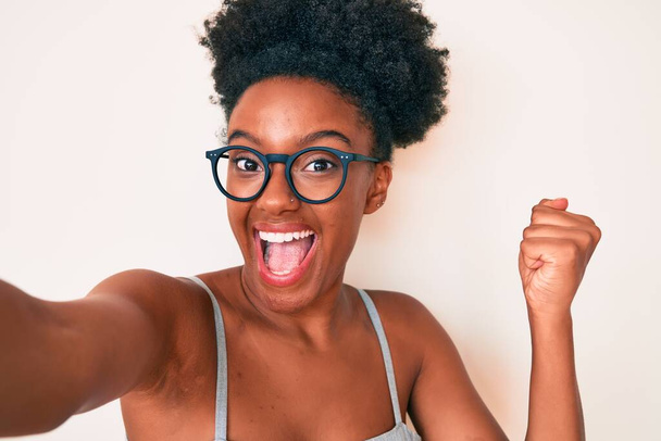 Mladá afroameričanka nosí ležérní oblečení a brýle, takže selfie u kamery křičí hrdě, slaví vítězství a úspěch velmi vzrušený se zvednutými pažemi  - Fotografie, Obrázek