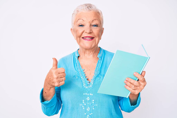 Senior beautiful woman with blue eyes and grey hair reading a book smiling happy and positive, thumb up doing excellent and approval sign  - 写真・画像