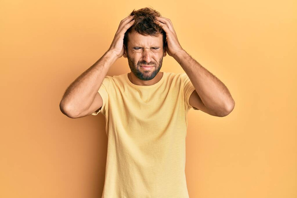 Hombre guapo con barba llevando camiseta amarilla casual sobre fondo amarillo que sufre de dolor de cabeza desesperado y estresado porque el dolor y la migraña. manos en la cabeza.  - Foto, Imagen