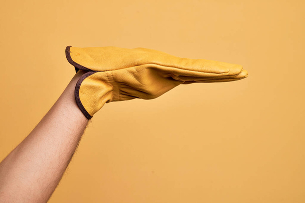 Hand of caucasian young man with gardener glove over isolated yellow background with flat palm presenting product, offer and giving gesture, blank copy space - Photo, Image