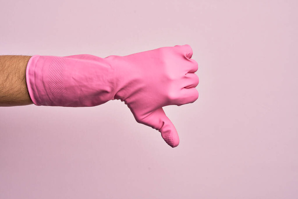 Mano de joven caucásico con guante de limpieza sobre fondo rosa aislado haciendo pulgares hacia abajo gesto de rechazo, desaprobación aversión y signo negativo - Foto, imagen