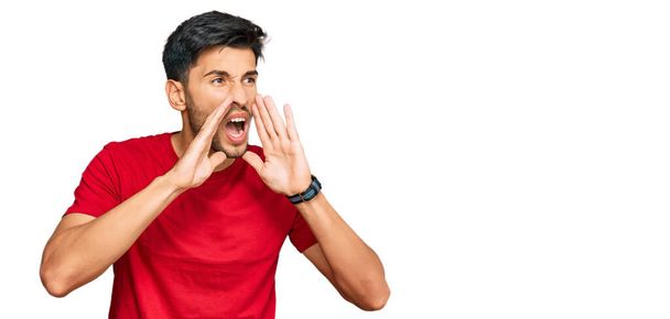 Young handsome man wearing casual red tshirt shouting angry out loud with hands over mouth  - Fotoğraf, Görsel