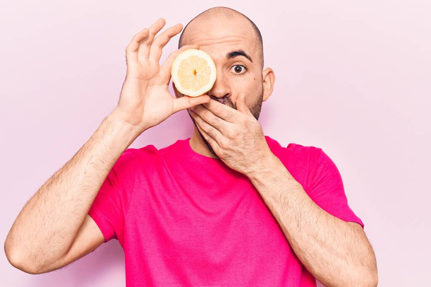 Mladý pohledný plešatý muž, držící rukou kousek citronu přes oko, zakrývající ústa, šokovaný a vystrašený z omylu. překvapený výraz  - Fotografie, Obrázek