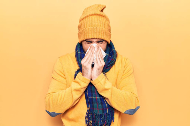 Young man illness using paper handkerchief on nose. Standing wearing scarf and wool cap over isoltated yellow background - Фото, изображение