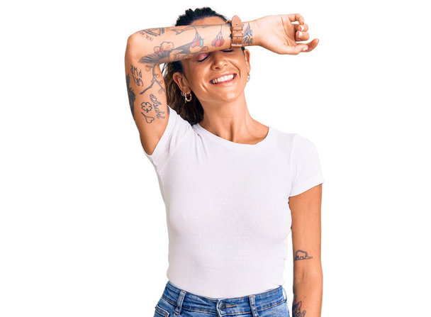 Young hispanic woman with tattoo wearing casual white tshirt covering eyes with arm smiling cheerful and funny. blind concept.  - Photo, Image
