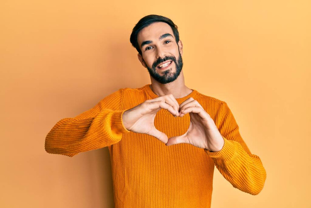 Joven hombre hispano vistiendo ropa casual sonriendo en amor haciendo forma de símbolo del corazón con las manos. concepto romántico.  - Foto, Imagen