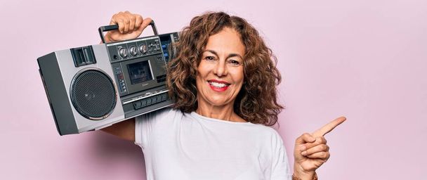 Middelbare leeftijd brunette hipster vrouw met retro muziek boombox over roze geïsoleerde achtergrond glimlachen gelukkig wijzend met hand en vinger naar de zijkant - Foto, afbeelding