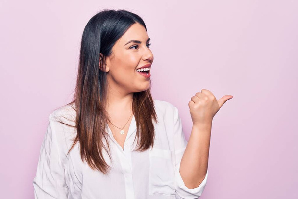 Young beautiful brunette woman wearing elegant shirt standing over isolated pink background pointing thumb up to the side smiling happy with open mouth - Photo, Image